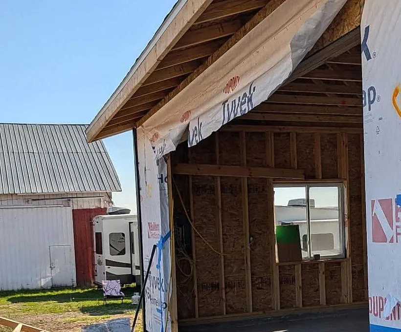 A newly framed pole barn wrapped in housewrap on a Northern Michigan property, an older white barn with a red door and a parked travel trailer standing behind it.