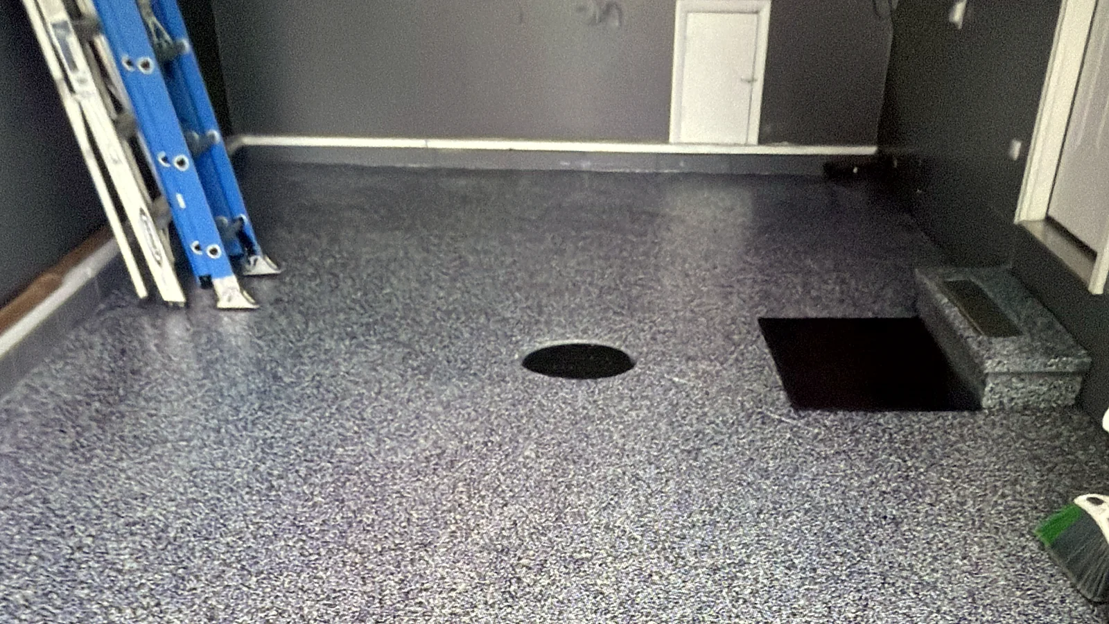 Finished blue-grey flake epoxy floor across a garage, run tight to the wall and cut cleanly around the floor drain, with a step and a ladder at the edges.