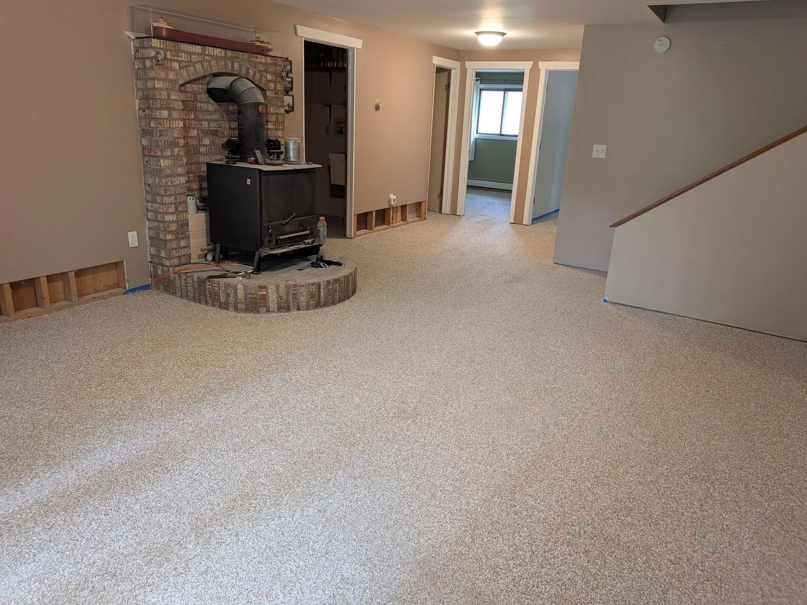 Finished beige flake epoxy floor across a living room, wood stove on a brick hearth to the right and a hallway beyond.