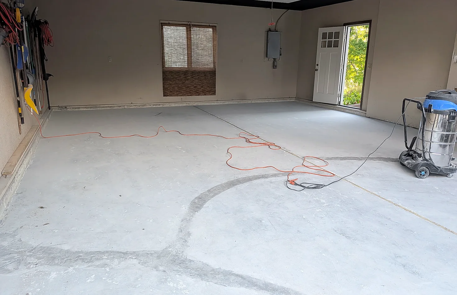 A two-car garage with the concrete ground back to bare grey, extension cord and dust-extraction vacuum still on the slab, ready for coating.