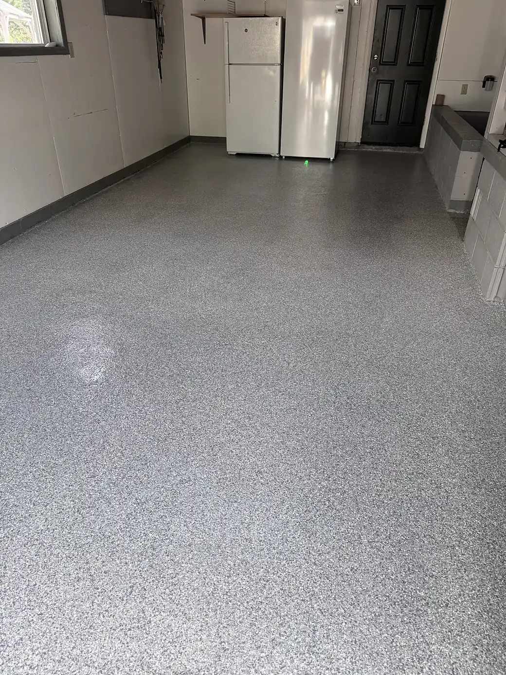 A finished grey flake epoxy floor running the length of a residential garage, past two upright freezers to a black side door.