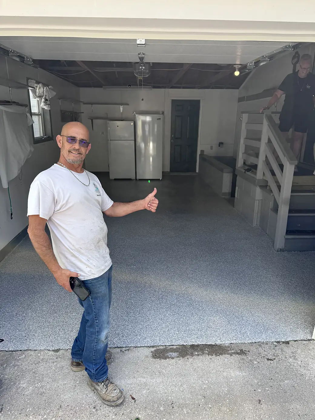 A man in a white T-shirt giving a thumbs up at the open door of the finished garage, the homeowner standing on the steps behind him.