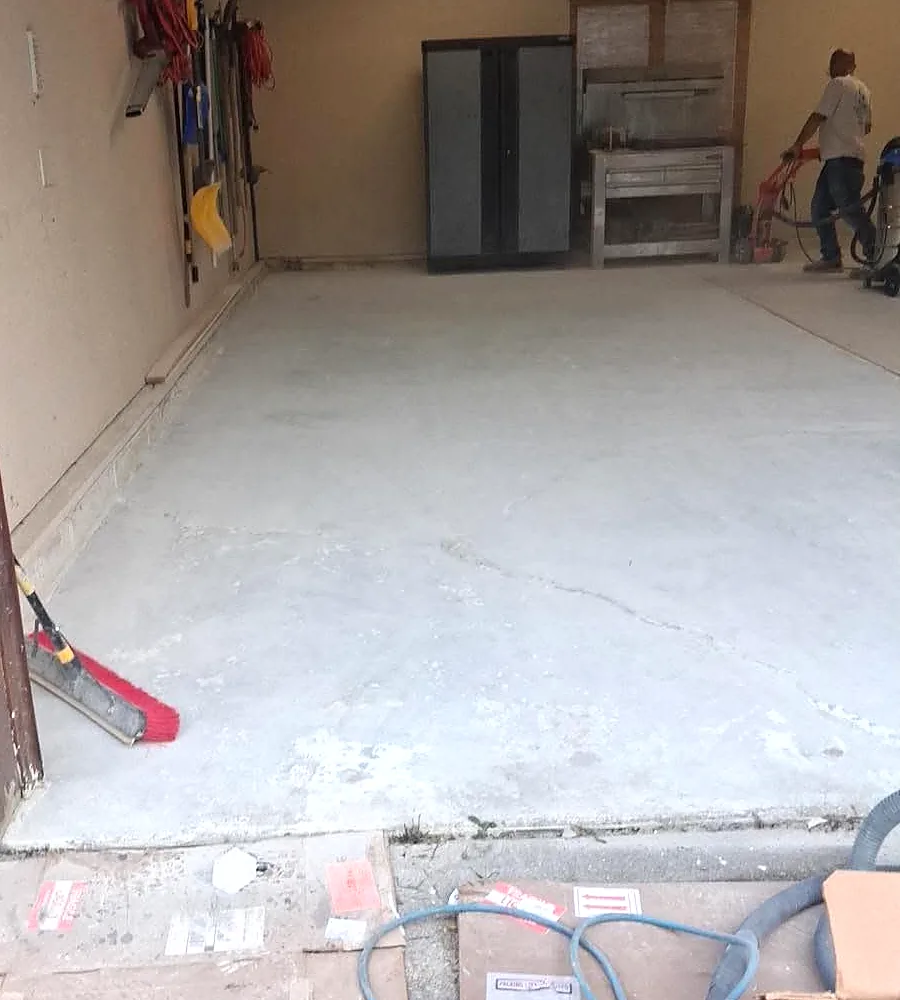 A bare, stained garage slab part-way through mechanical preparation, with a floor scraper and a dust-extraction hose laid across the concrete.
